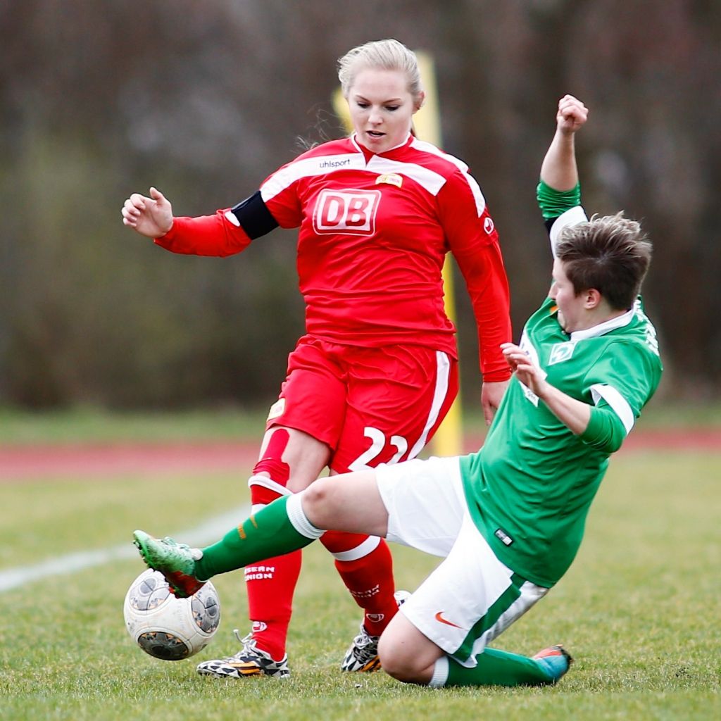 Zwei Frauen im Fußballtrikot, eine in Rot und die andere in Grün, kämpfen um den Ball auf einem Rasenplatz.
