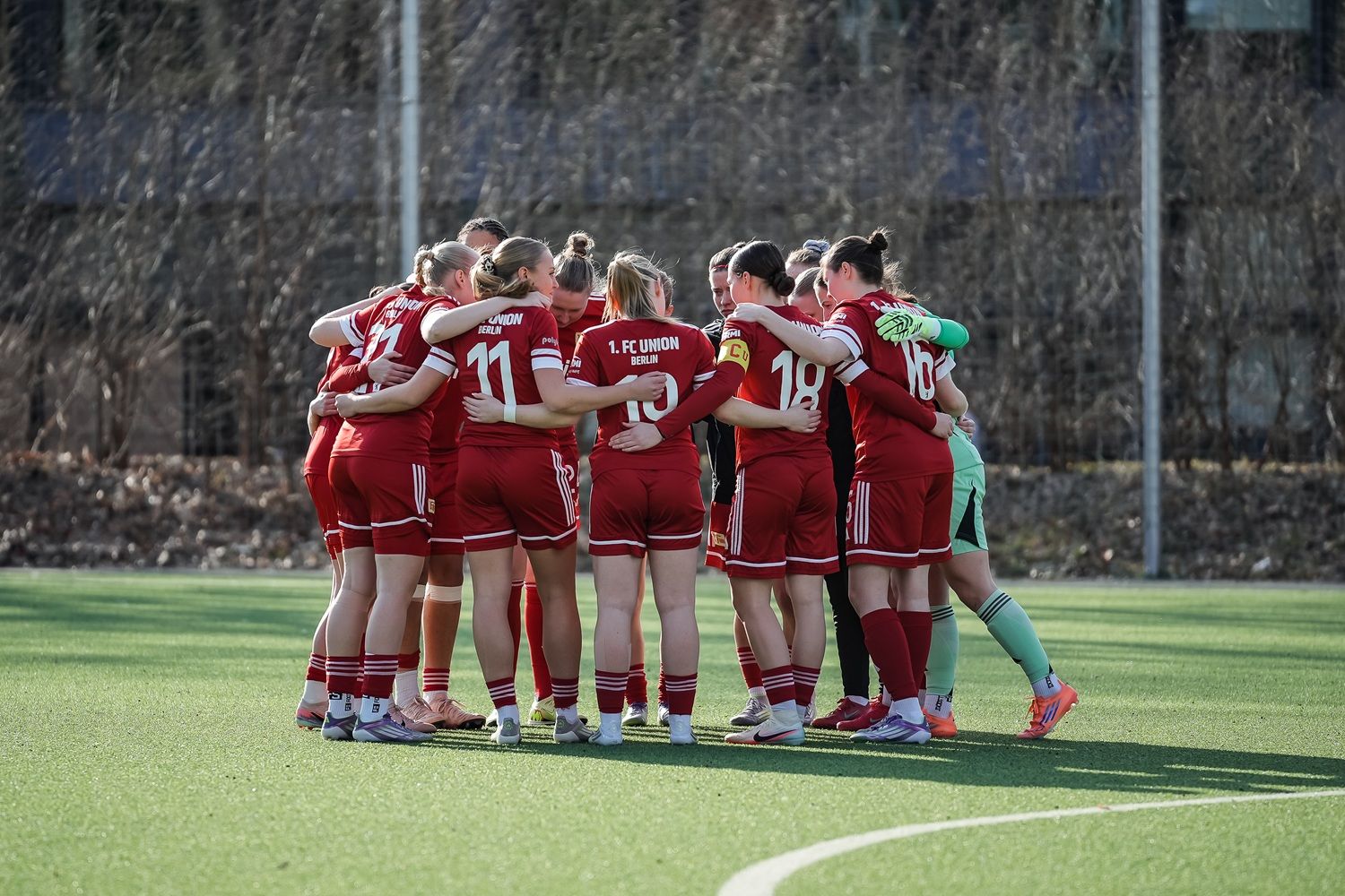 Mannschaft in roten Trikots steht im Kreis zusammen und hält sich an den Schultern, vor einem Fußballspiel auf einem Rasenplatz.