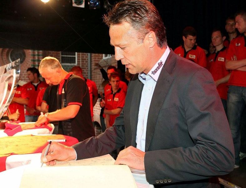 Ein Mann in Anzug signiert ein Dokument, während im Hintergrund weitere Personen in roten T-Shirts stehen.