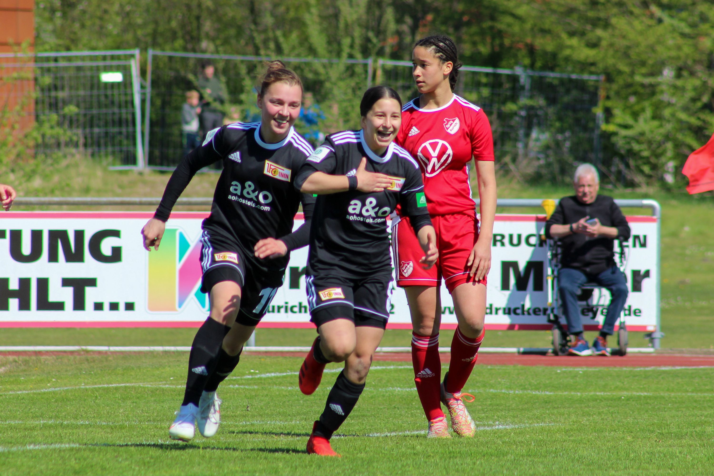 Zwei Spielerinnen in schwarz-weißen Trikots feiern ein Tor, während eine Spielerin in rot passiv im Hintergrund steht.