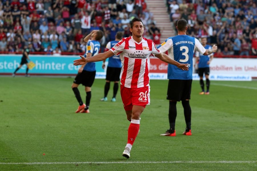 A football player in a red and white uniform celebrates after a goal, while other players react in the background.
