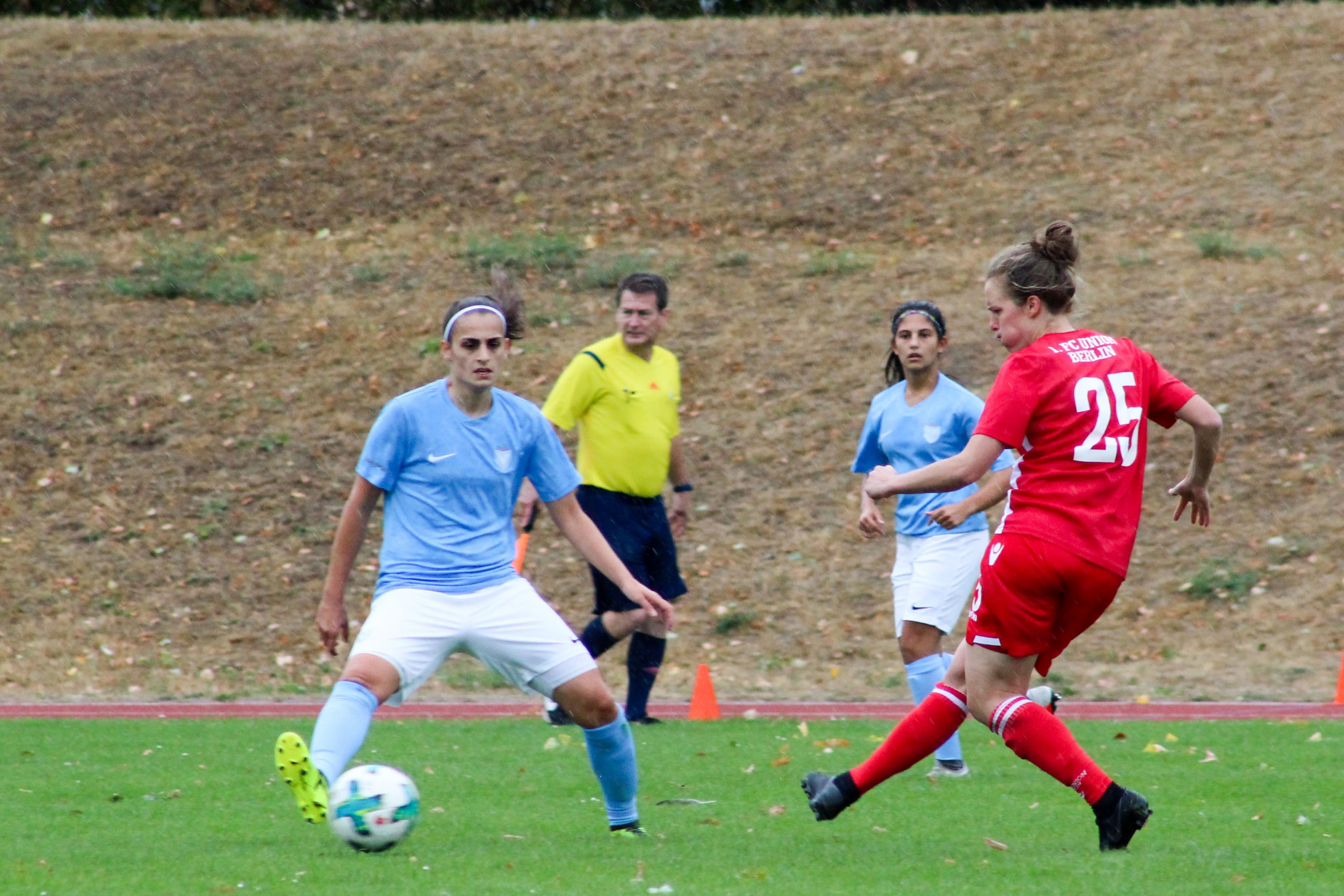 Spielszene eines Fußballspiels mit Spielerinnen in roten und blauen Trikots, während eine Spielerin den Ball dreht. Schiedsrichter im Hintergrund.