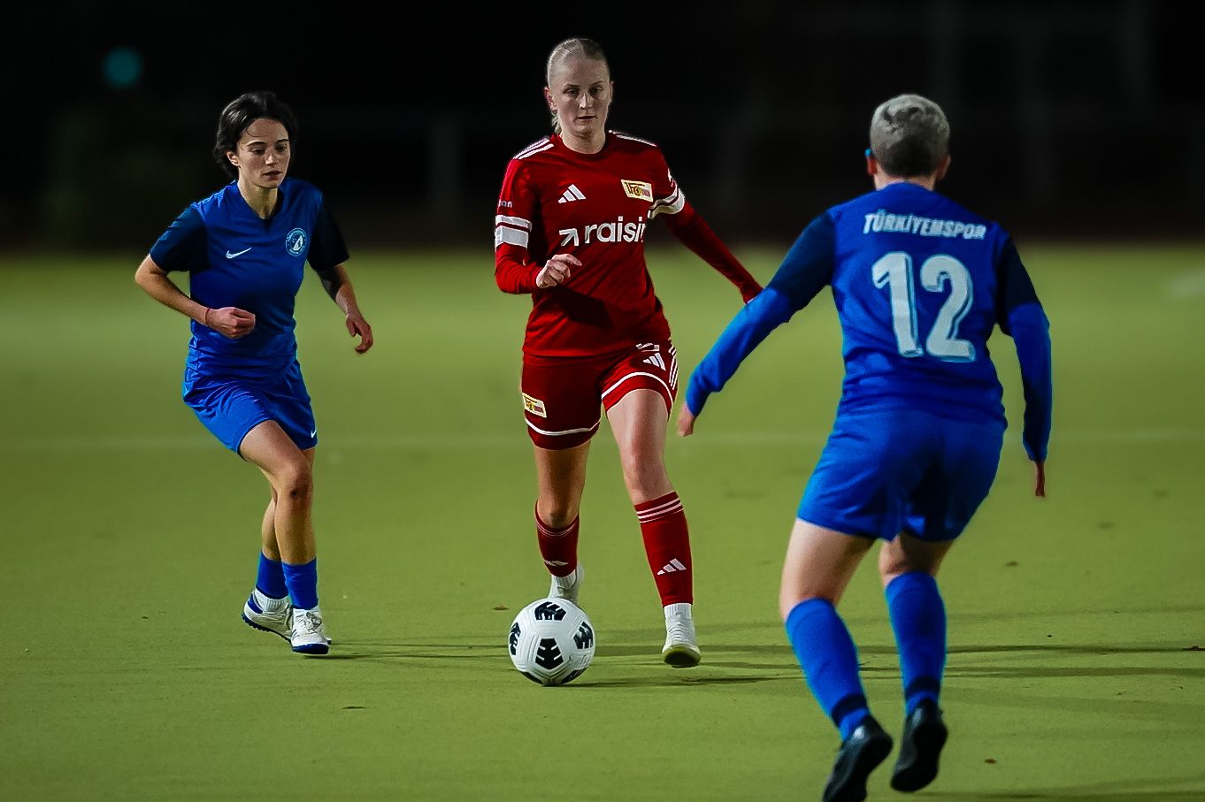 Eine Fußballspielerin in rotem Trikot dribbelt den Ball, während zwei Spielerinnen in blauen Trikots sie verteidigen.