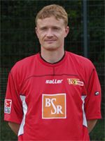 Ein Mann in einem roten Fußballtrikot steht vor einem Fußballfeld und schaut direkt in die Kamera.