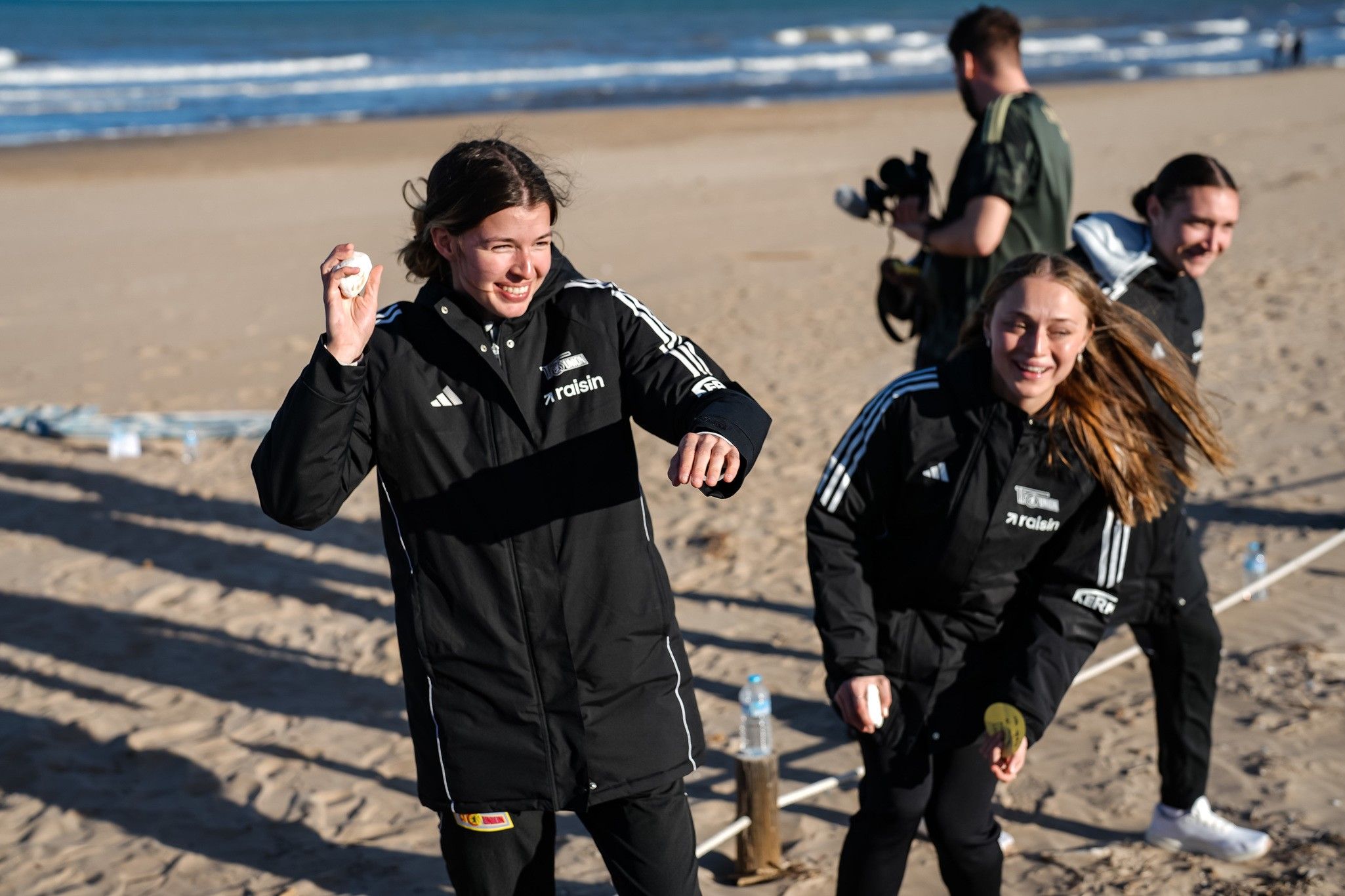 Zwei Frauen in schwarzen Jacken lachen und werfen einen Ball am Strand, während im Hintergrund eine weitere Person und eine Wasserflasche sichtbar sind.