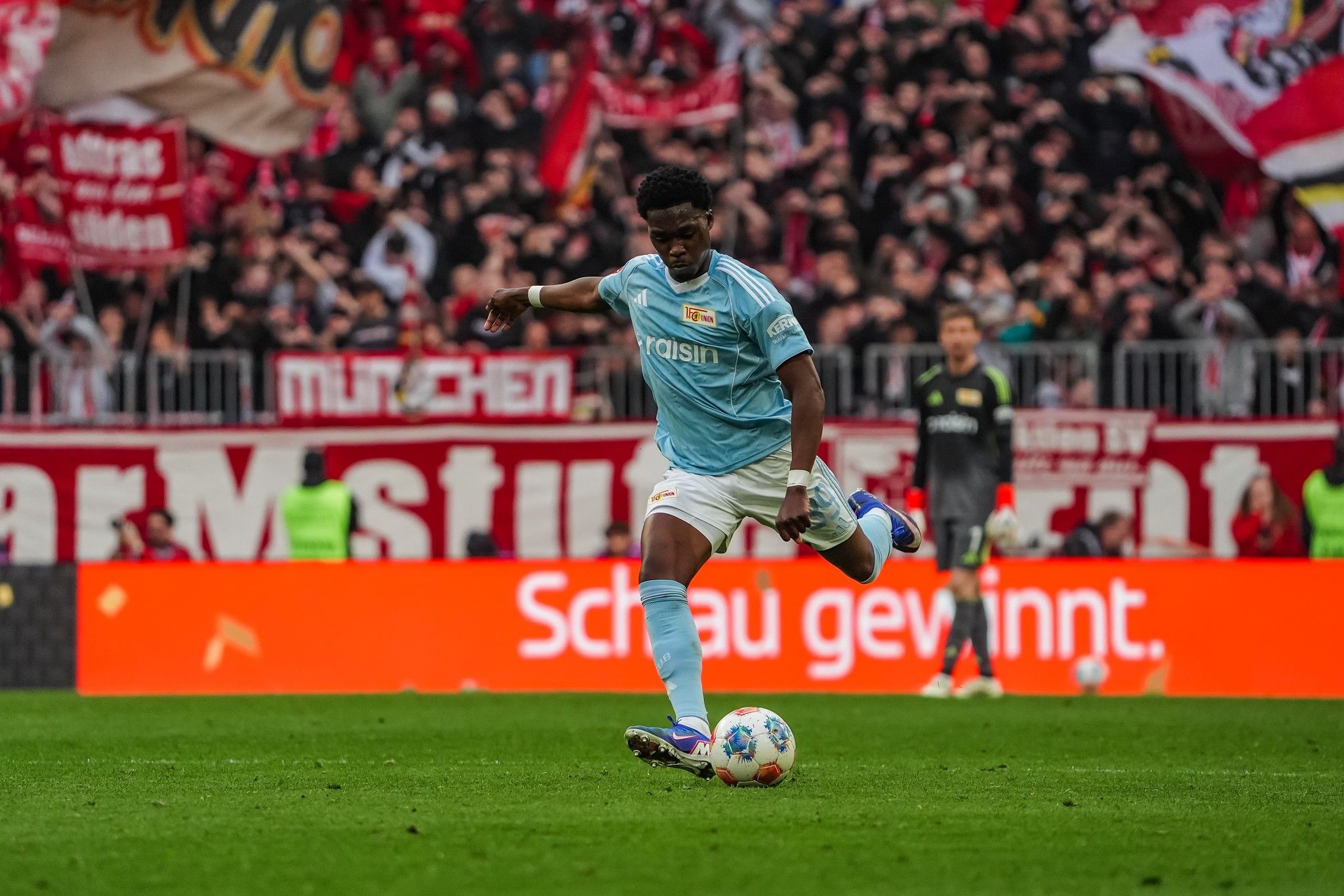 Torwart in blauer Trikotausführung, der einen Fußball im Stadion schießt, umgeben von jubelnden Fans im Hintergrund.