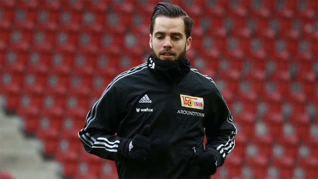 A young man in sportswear jogs on a football field with red stands in the background.
