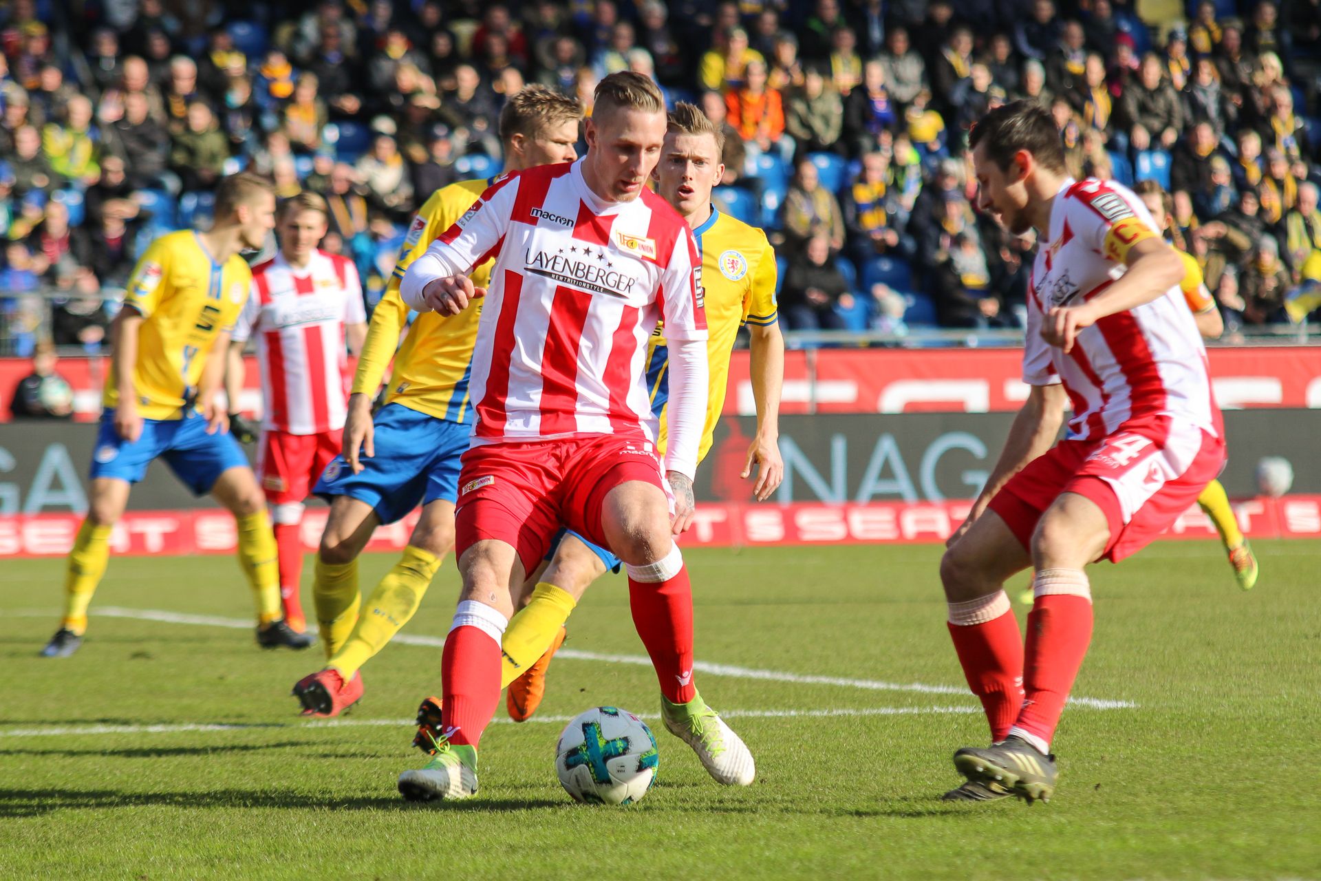 Zwei Fußballspieler in roten und weißen Trikots dribbeln den Ball, während ein Spieler in gelbem Trikot zuschaut. Zuschauer im Hintergrund.