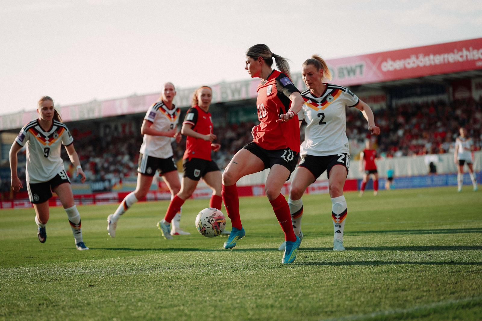 Eine Fußballspielszene mit Spielerinnen in roten und weißen Trikots, die um den Ball kämpfen. Zuschauer im Hintergrund.
