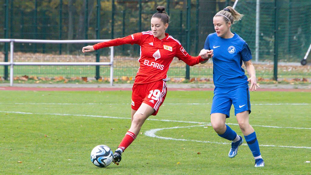 Zwei Fußballspielerinnen in Aktion auf einem Spielfeld, eine in rotem Trikot und die andere in blauem Trikot.
