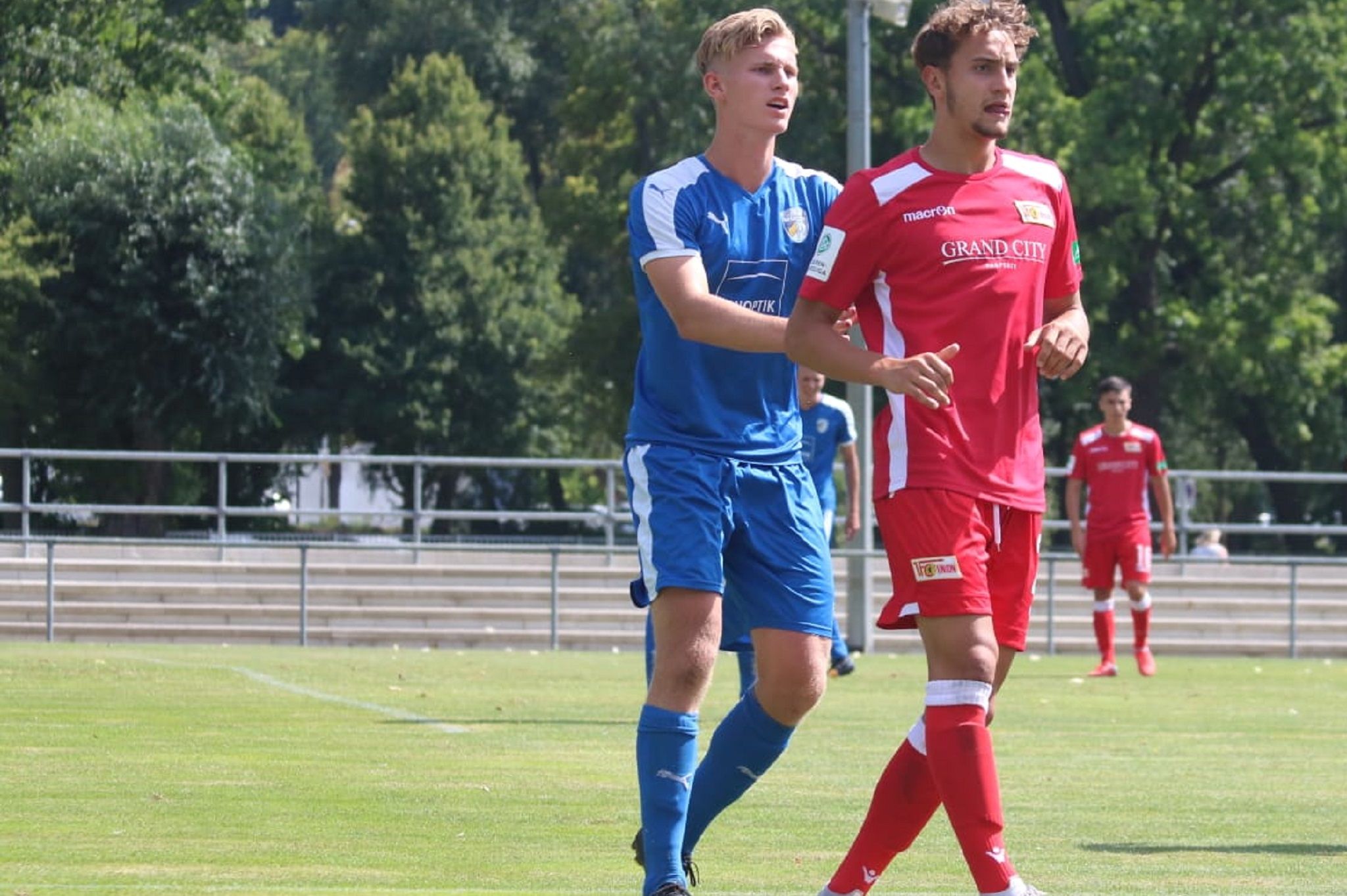 Zwei Fußballspieler auf einem Rasenplatz, einer in blauer und der andere in roter Uniform, interagieren während des Spiels.