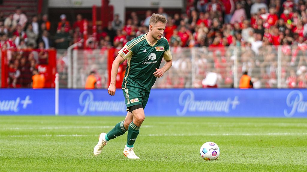 Ein Fußballspieler in grüner Uniform läuft auf dem Spielfeld, umgeben von jubelnden Fans im Hintergrund.