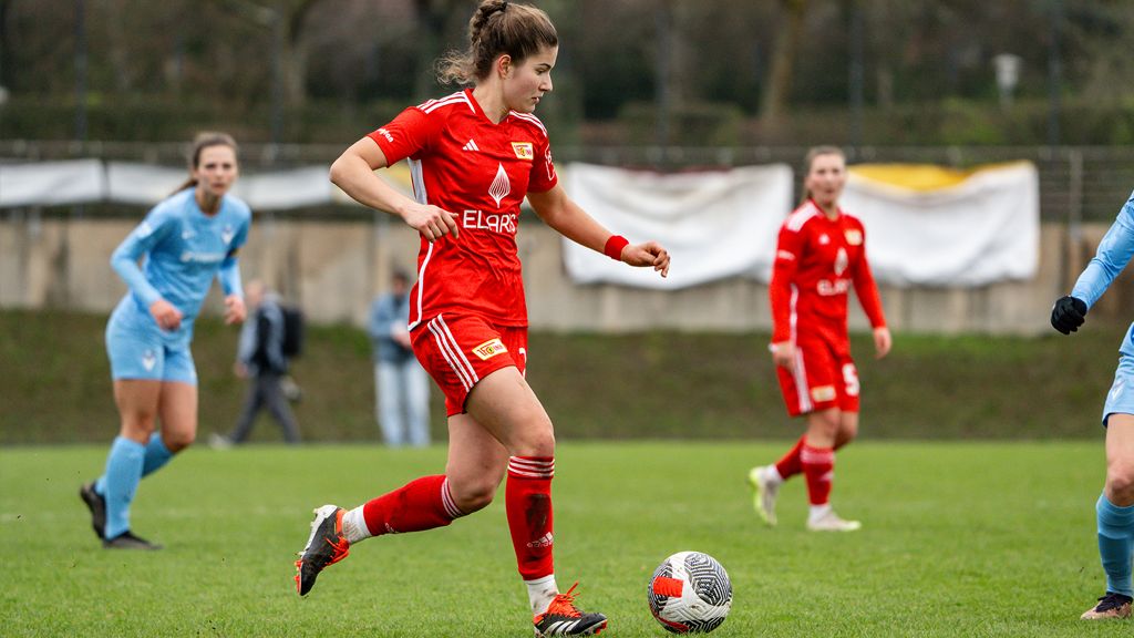 Eine Fußballspielerin in rotem Trikot dribbelt den Ball auf dem Spielfeld, während andere Spielerinnen im Hintergrund stehen.