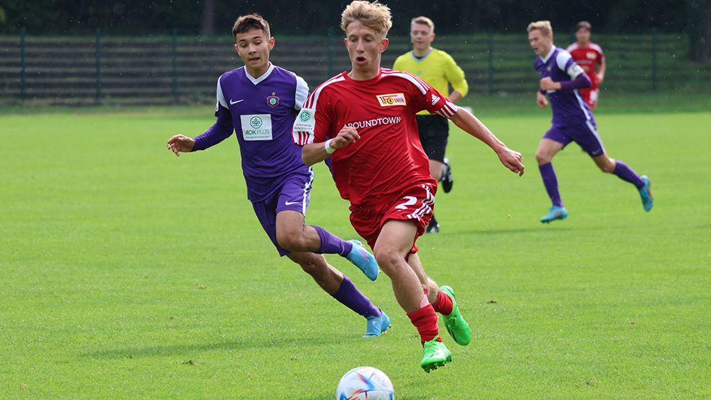 Ein junger Fußballspieler in rotem Trikot sprintet mit dem Ball, während ein Gegner in lila Trikot ihn verfolgt.