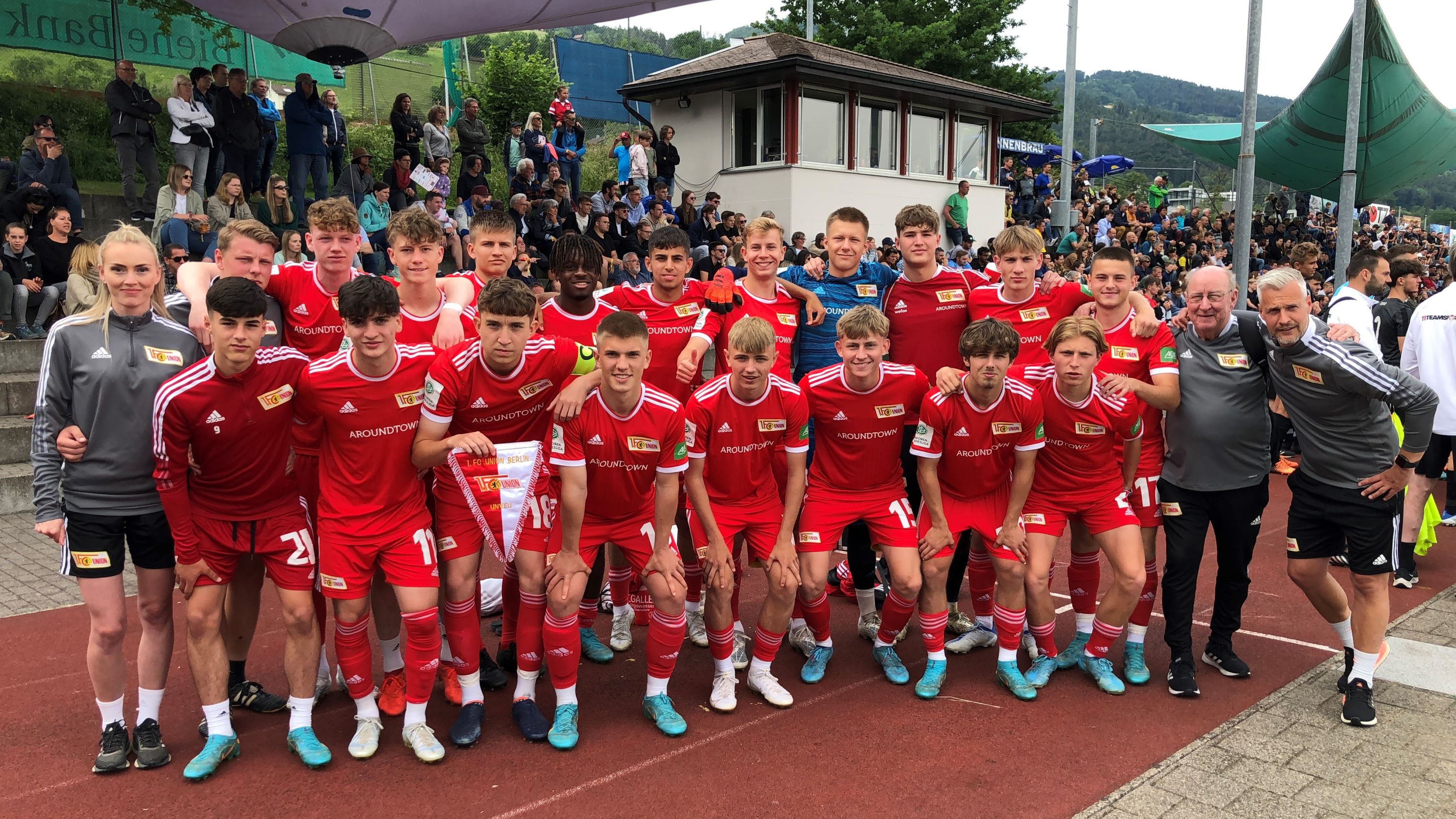Eine Gruppe von 18 Fußballspielern in roten Trikots posiert gemeinsam an einem Stadionrand. Im Hintergrund sind Zuschauer zu sehen.