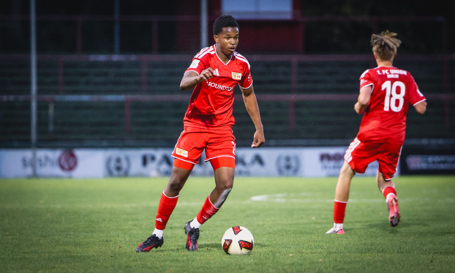 Ein Fußballspieler in rotem Trikot dribbelt den Ball auf einem grünen Spielfeld, während ein Mitspieler in der Nähe ist.