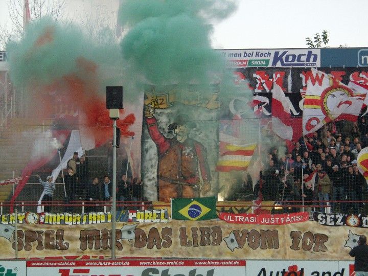 Rauchtore in den Vereinsfarben und Banner von Fans in einem Stadion, während eine Feierlichkeit oder ein Support-Event stattfindet.