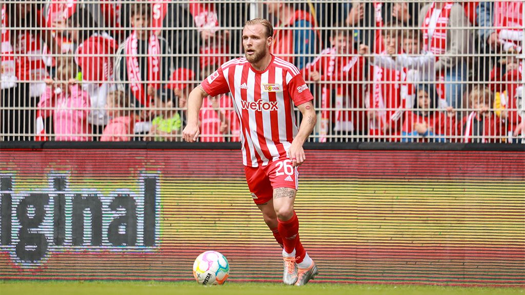 A football player in red and white stripes runs across the field while many fans cheer in the background.