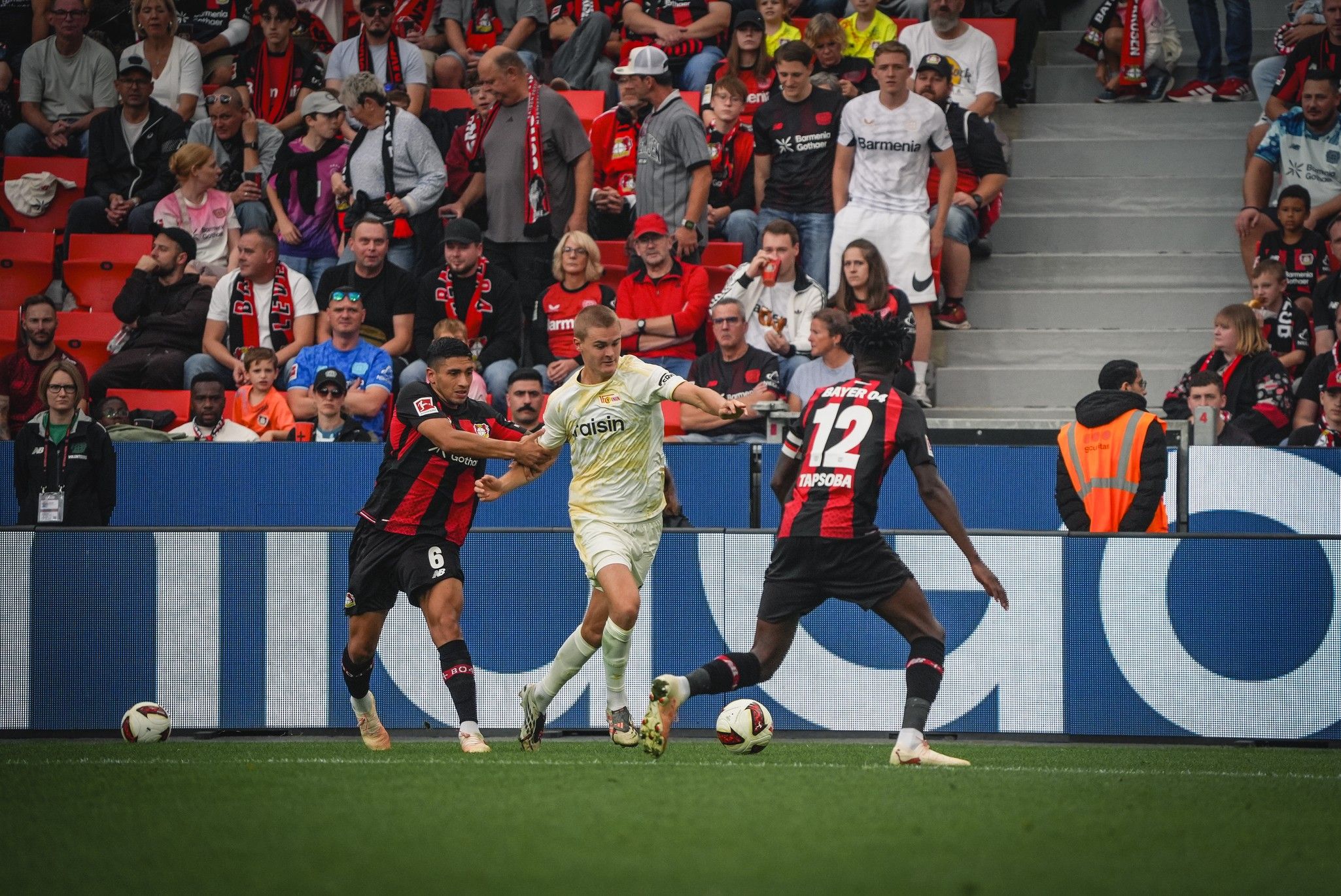 Zwei Fußballspieler versuchen, den Ball von einem dritten Spieler zu erobern, während die Fans im Hintergrund das Spiel verfolgen.