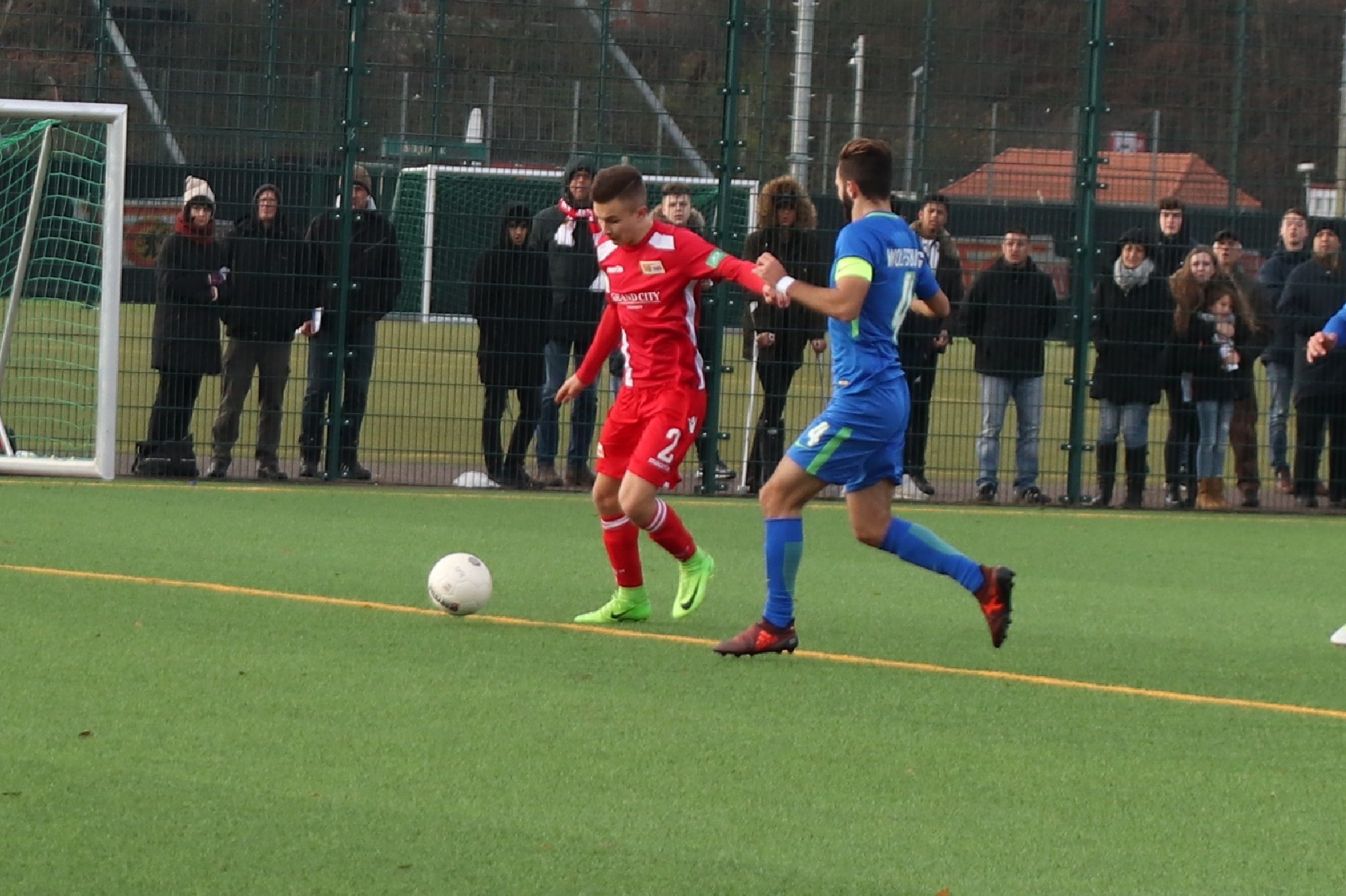 Ein Fußballspieler in roter Uniform dribbelt den Ball, während ein gegnerischer Spieler in blauer Uniform ihn verfolgt. Zuschauer am Rand.
