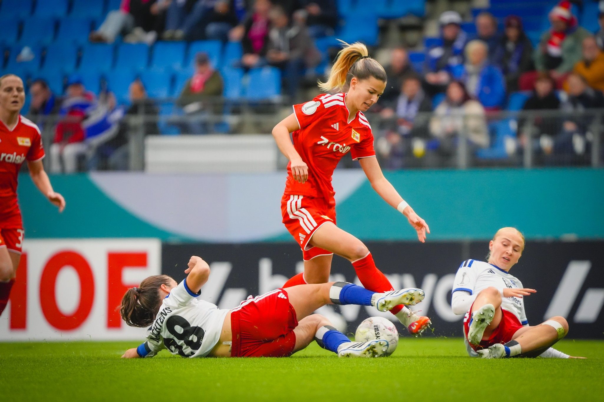Zwei Spielerinnen liegen auf dem Rasen, während eine dritte Spielerin in rotem Trikot an ihnen vorbeiläuft. Stadionhintergrund.