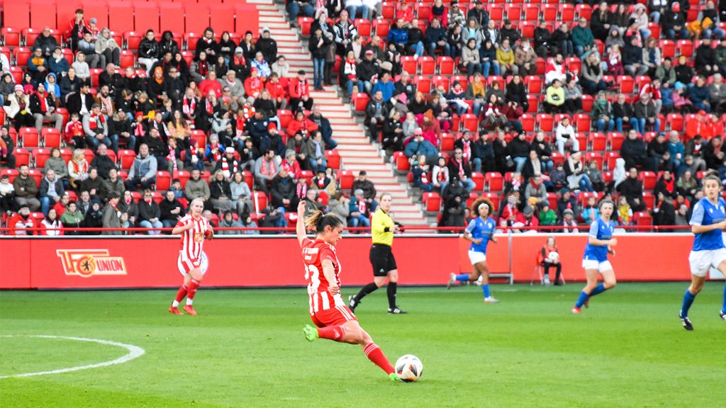 Game scene in a football stadium with players in red and blue jerseys, surrounded by a crowd in red seats.