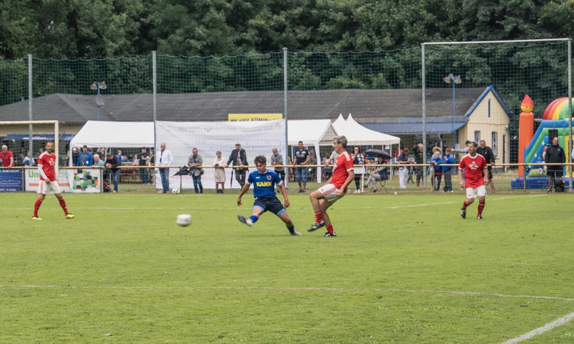 Ein Fußballspieler in blauer Trikotfarbe schießt auf das Tor, während zwei gegnerische Spieler in roten Trikots reagieren. Zuschauer im Hintergrund.