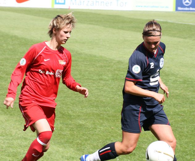 Zwei Frauen im Fußballtrikot spielen auf einem Rasenfeld, eine dribbelt den Ball, die andere verfolgt sie.
