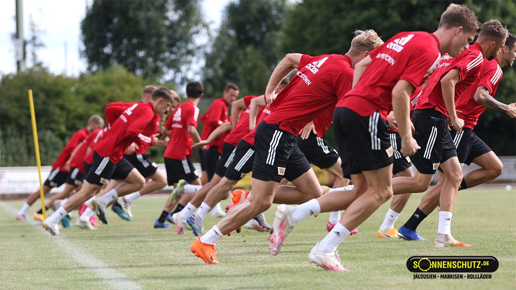A group of soccer players in red jerseys is sprinting on a training field.
