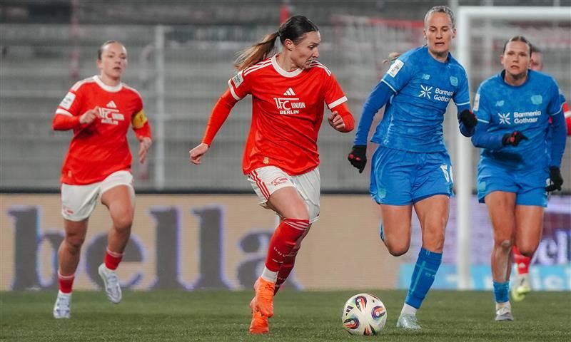 Eine Spielerin in rotem Trikot dribbelt den Ball, während zwei Gegnerinnen sie verteidigen. Im Hintergrund sind weitere Spielerinnen zu sehen.