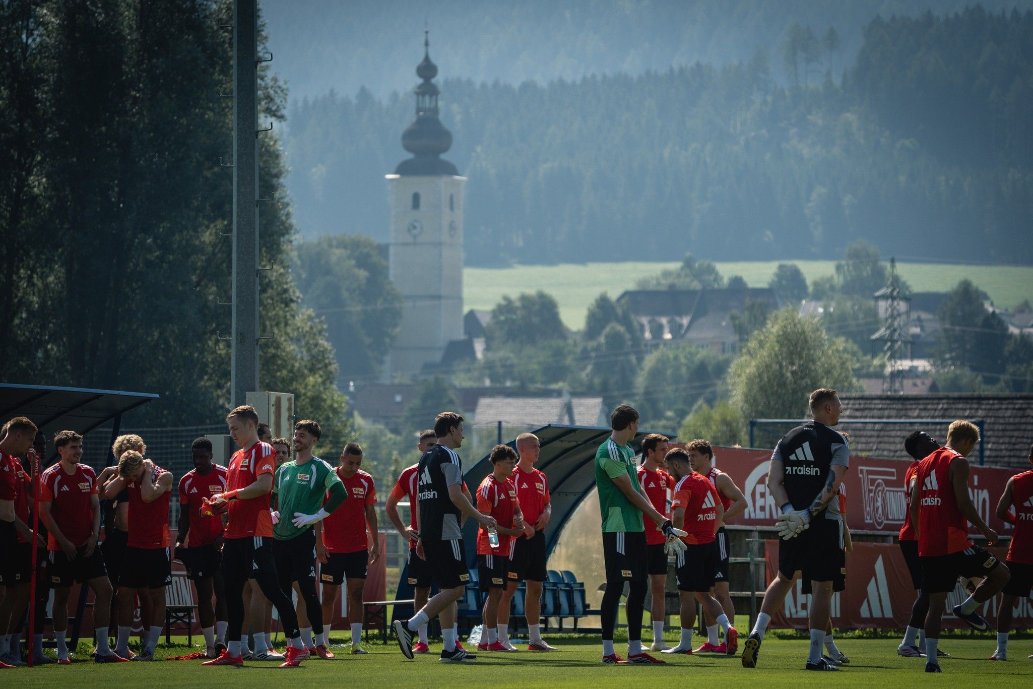 Fußballmannschaft trainiert auf einem grünen Platz, im Hintergrund eine Kirche und bewaldete Hügel.