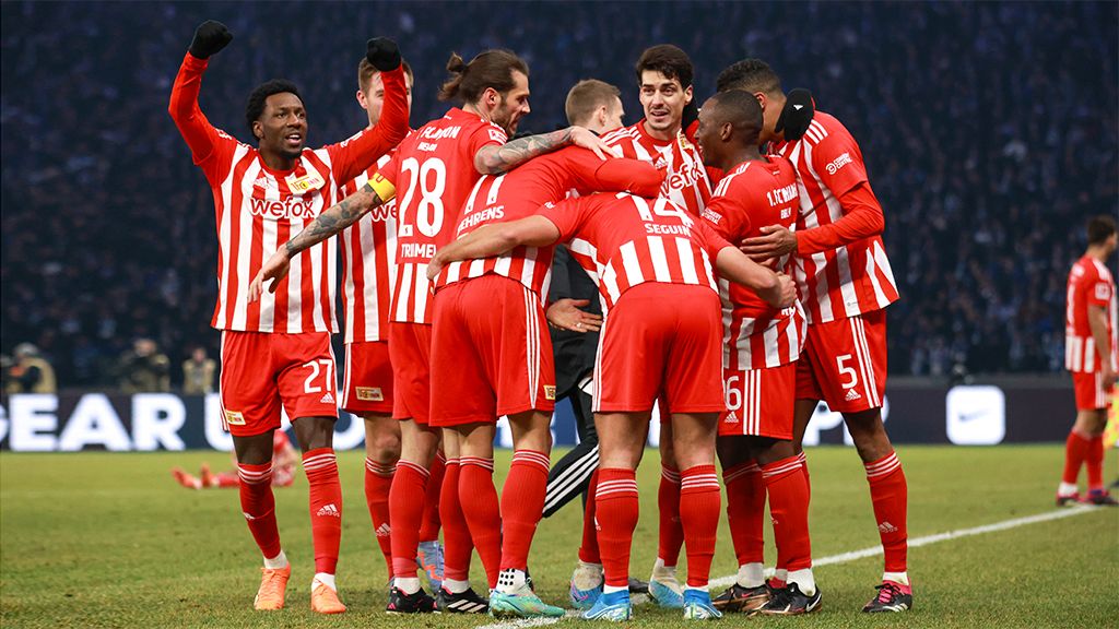 A group of soccer players in red jerseys joyfully embraces on the field.