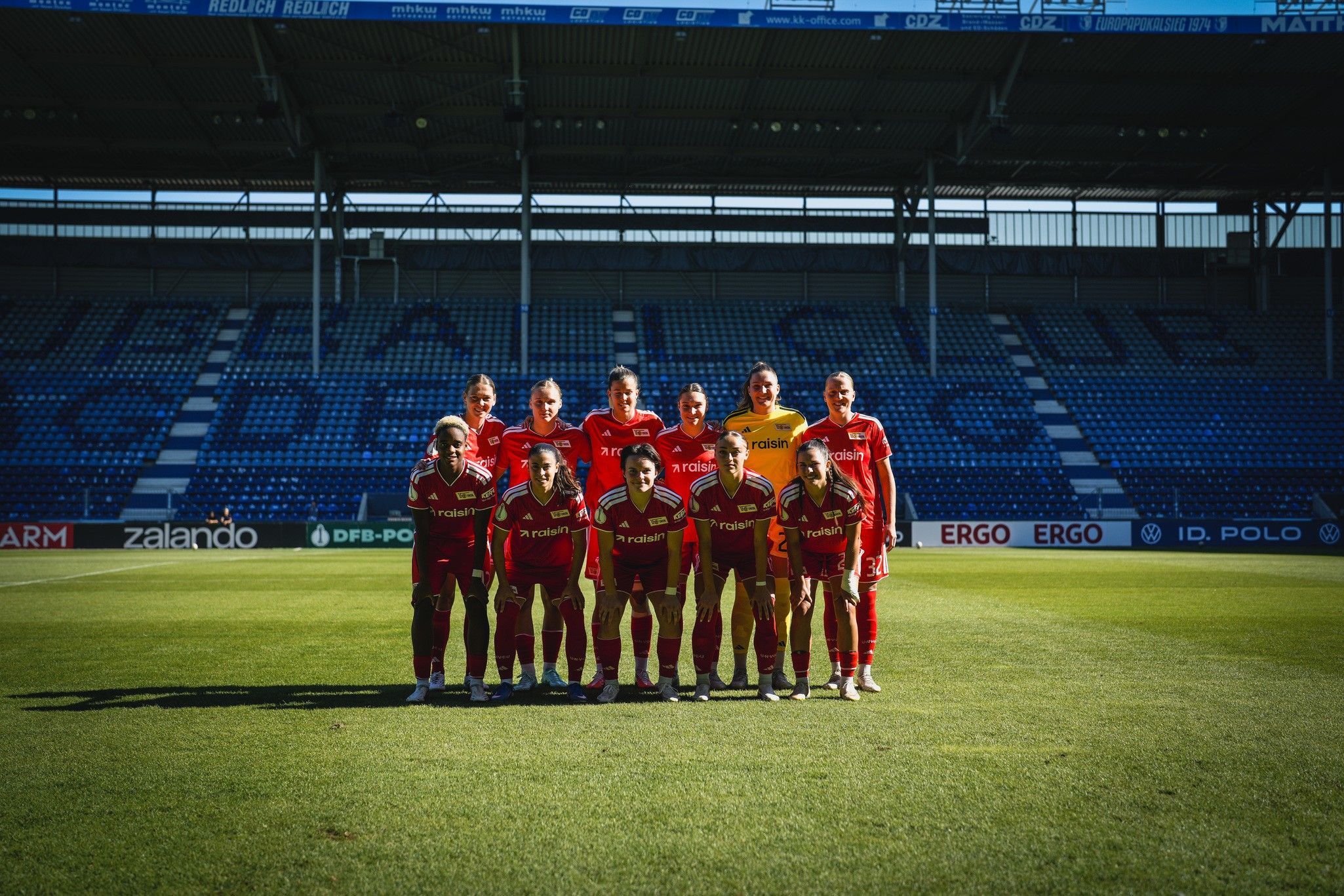 Eine Gruppe von zehn Fußballspielern in roten Trikots posiert auf einem grünen Fußballfeld vor einer Tribüne.
