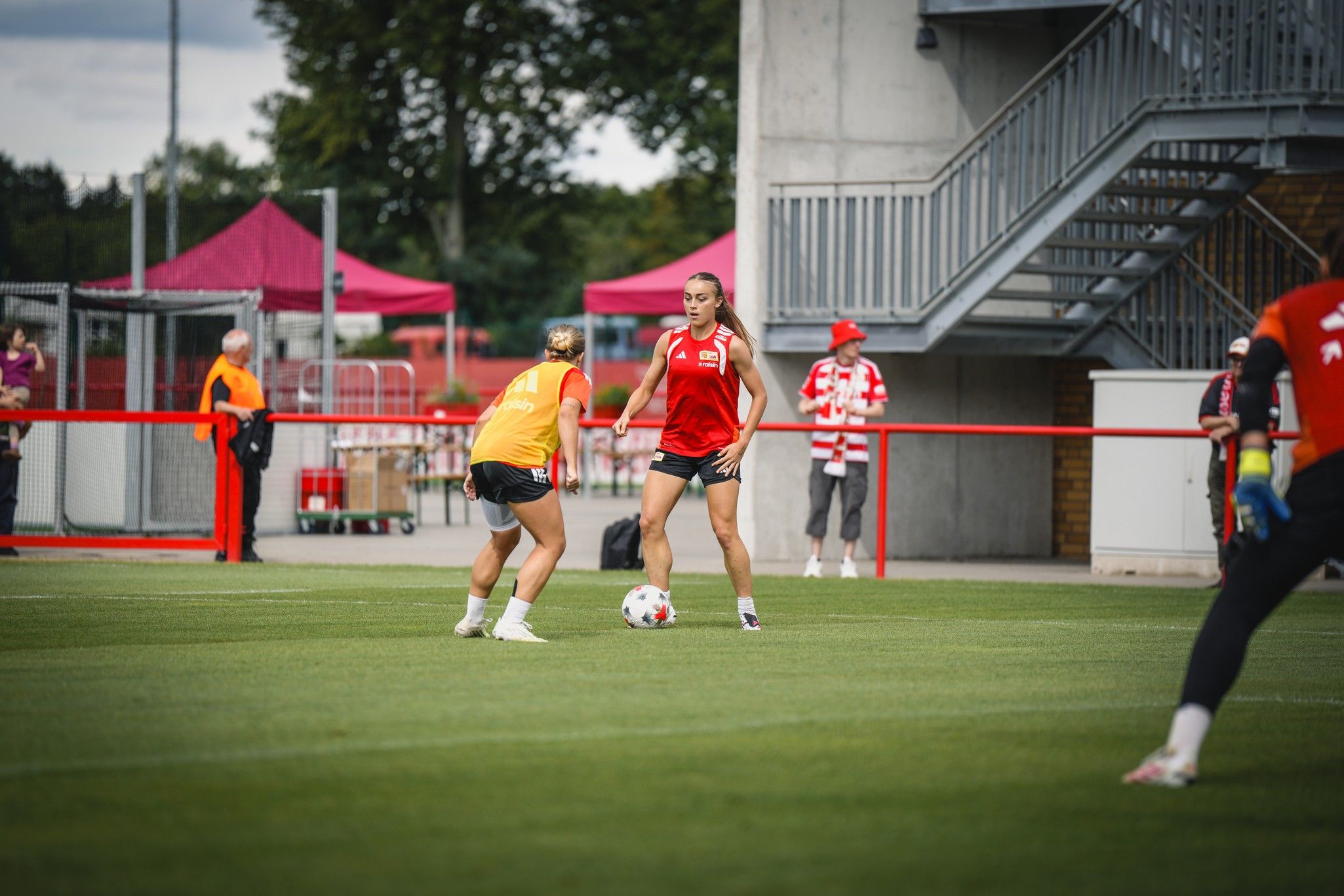 Zwei Fußballspielerinnen trainieren auf einem Rasenplatz, während Zuschauer im Hintergrund stehen.