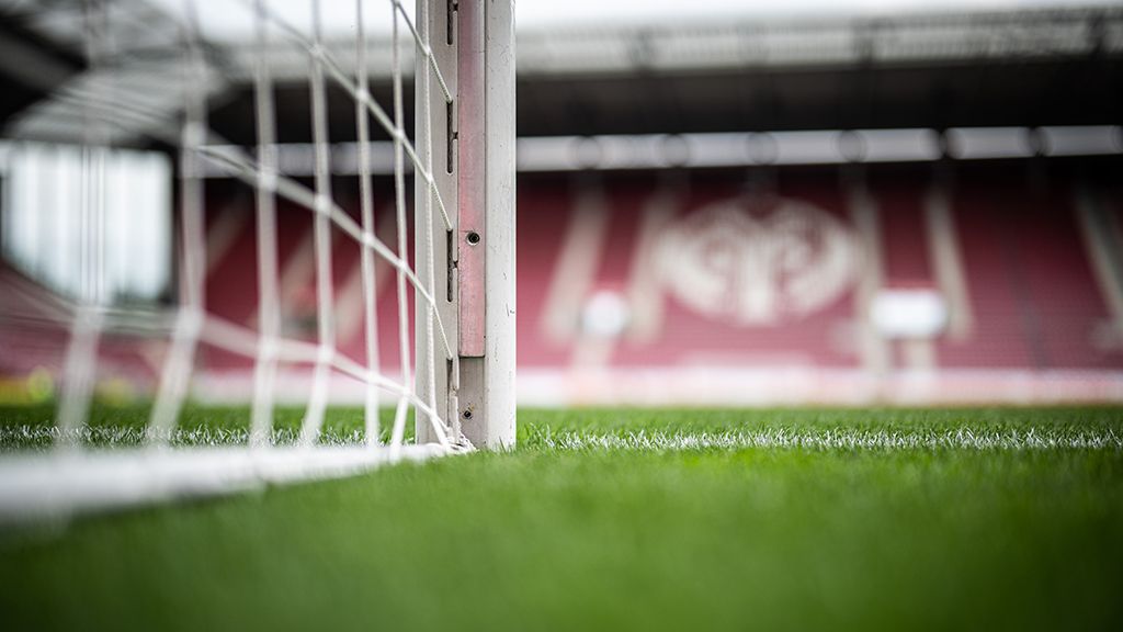 View through the goal net onto a green football field with stadium seating in the background.