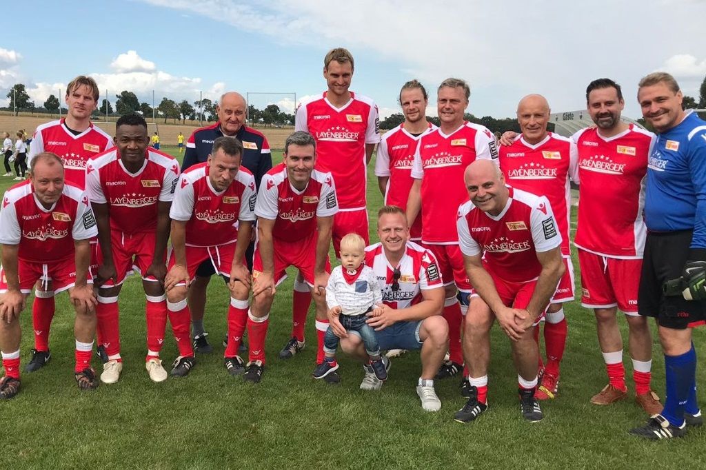 Eine Fußballmannschaft in roten Trikots posiert auf einem Rasenfeld, mit einem kleinen Jungen in der Mitte.