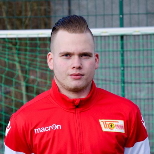 Ein junger Mann mit zurückgegeltem Haar trägt eine rote Sportjacke mit dem Logo von Union Berlin vor einem Fußballtor.