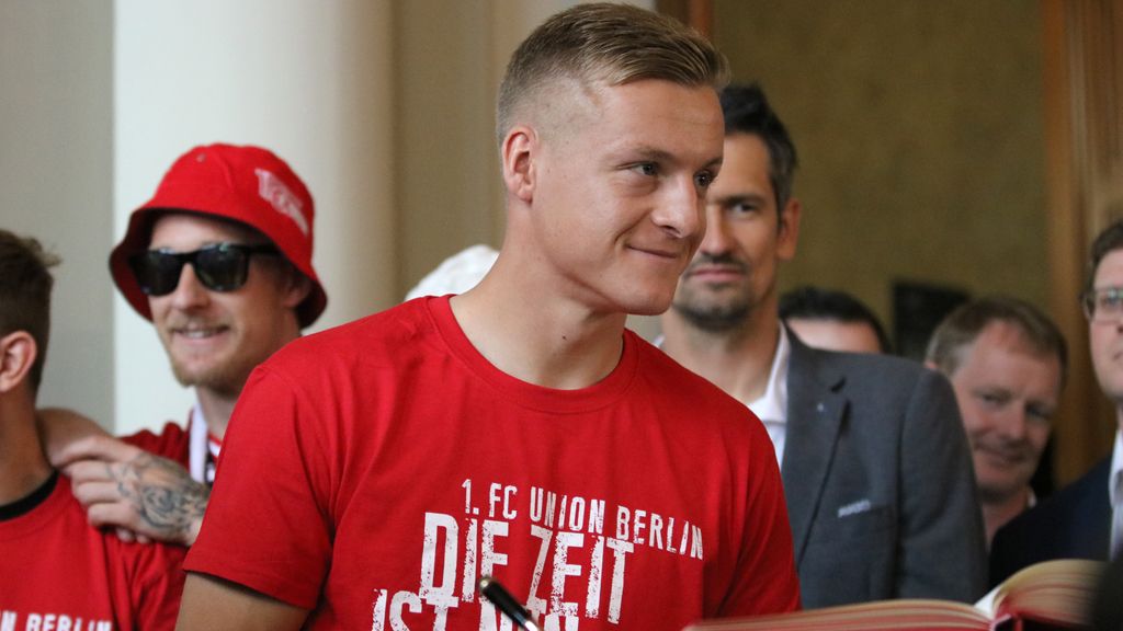 Ein junger Mann mit kurzem Haar und rotem T-Shirt steht lächelnd bei einer Veranstaltung, umgeben von weiteren Personen in roten Kleidungsstücken.