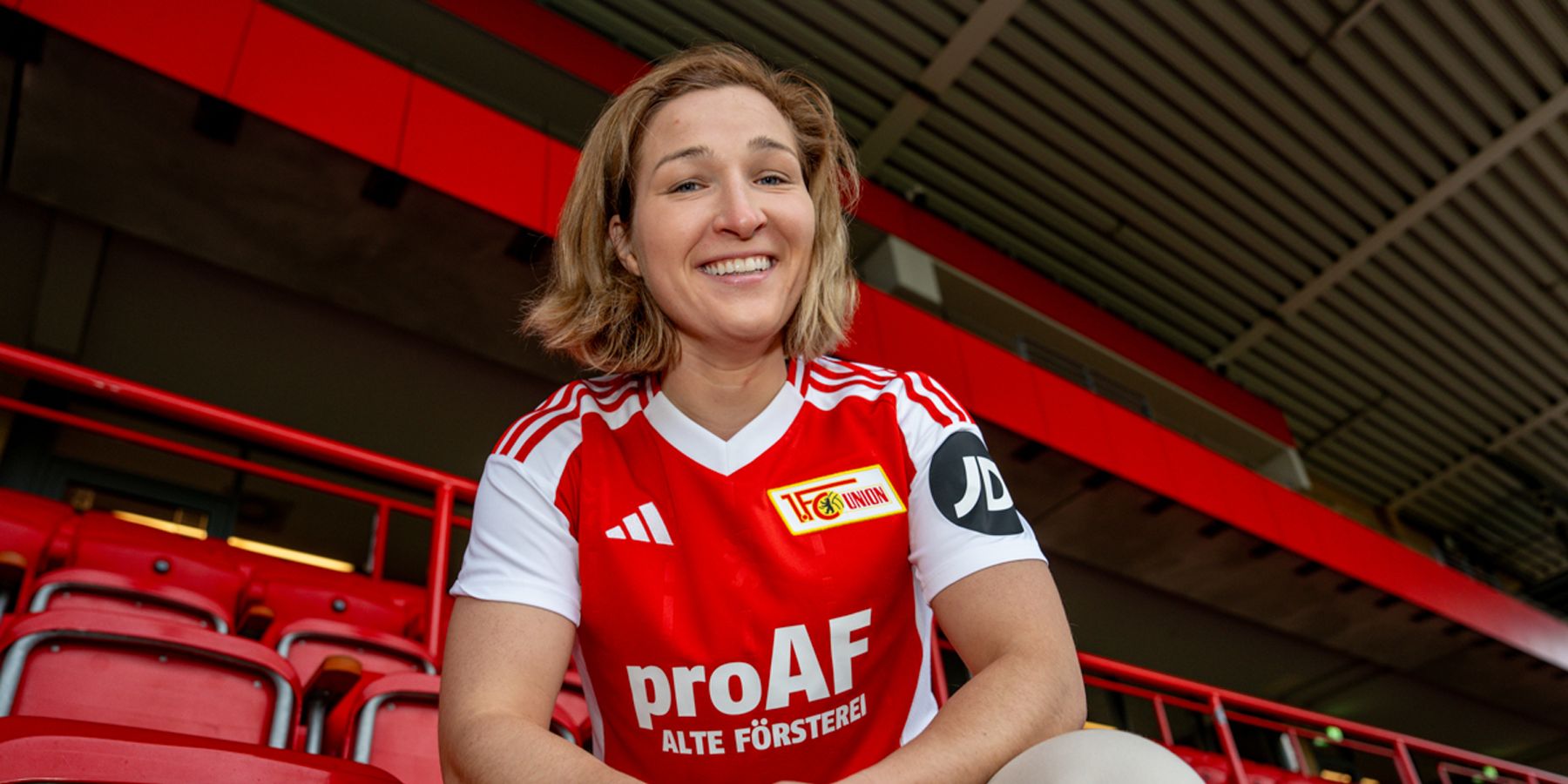 Eine lächelnde Person in einem roten Fußballtrikot sitzt auf einer Tribüne mit roten Sitzen im Hintergrund.