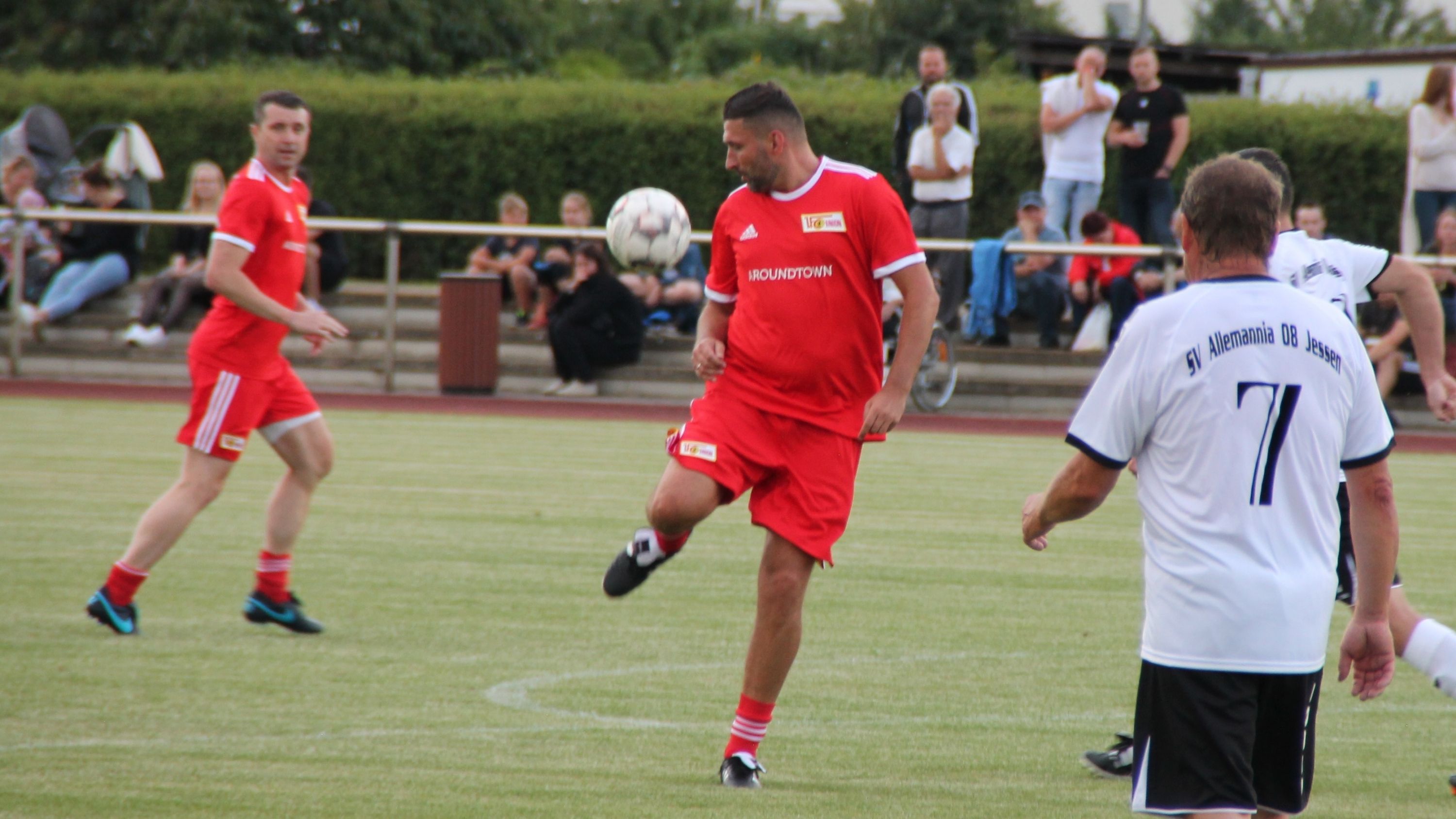 Spieler in roten Trikots kickt einen Fußball auf einem Rasenfeld, umgeben von Zuschauern.