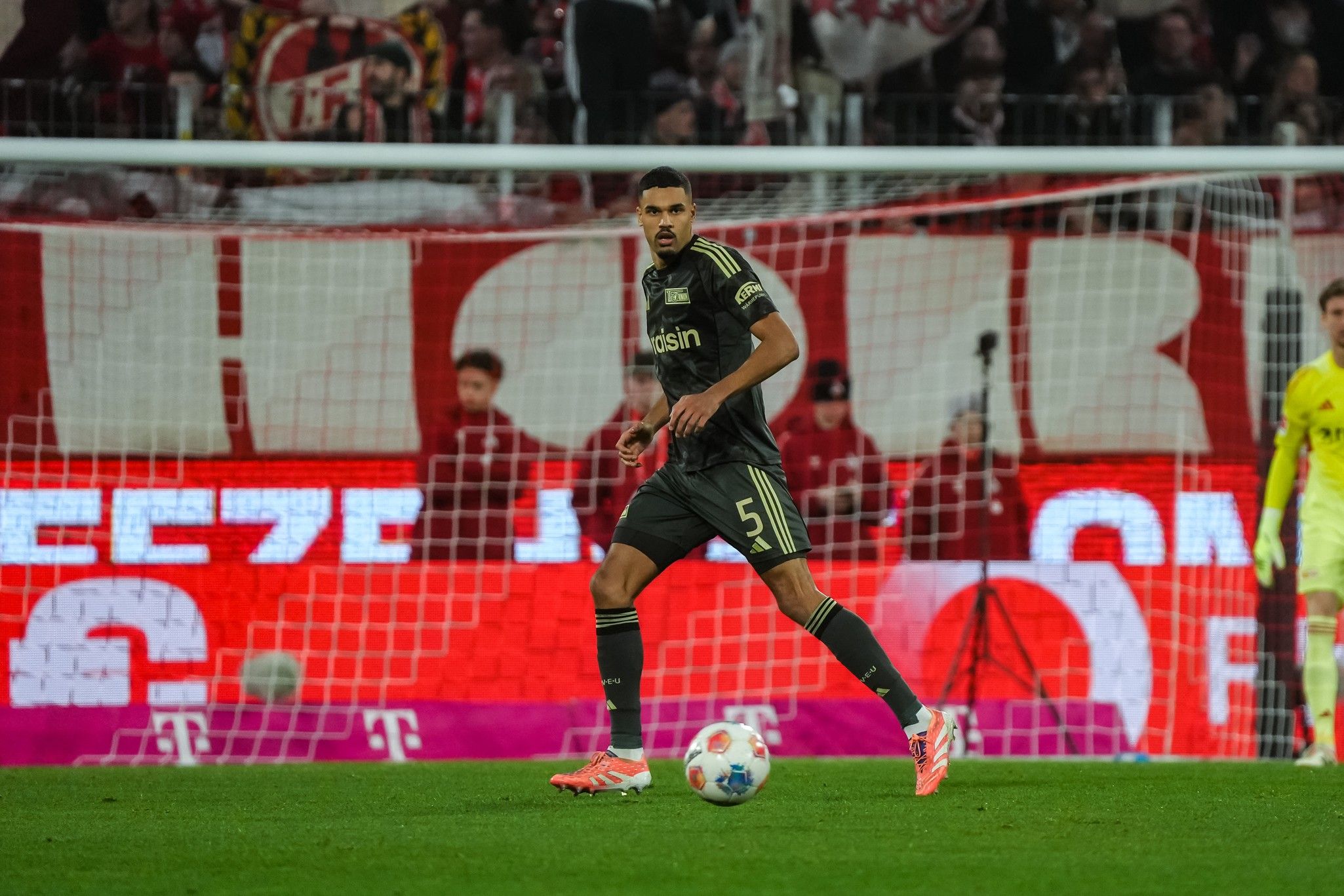 Ein Fußballspieler in einem schwarzen Trikot steht auf dem Spielfeld vor einem Tor, im Hintergrund Fans mit Schals und Banner.