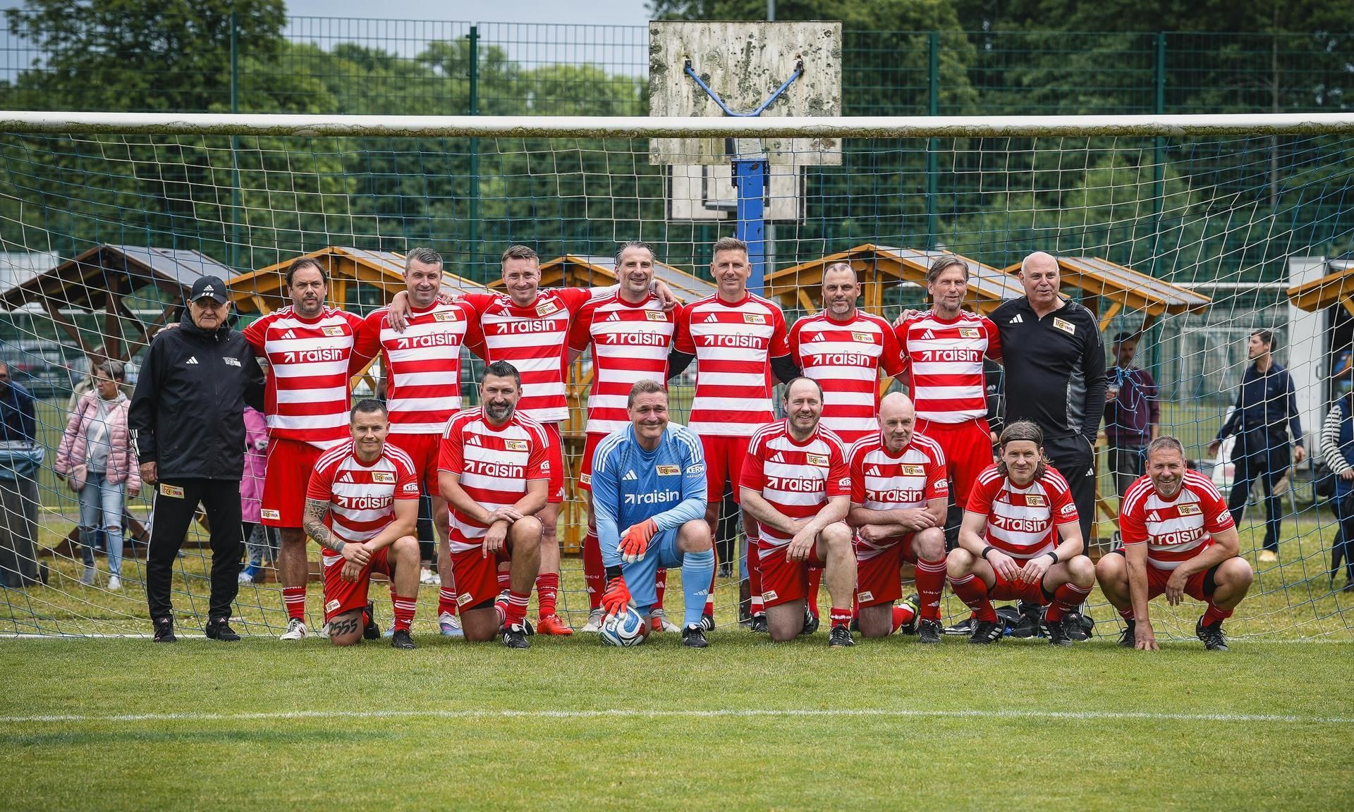 Eine Fußballmannschaft in roten Trikots steht vor einem Tor. Im Hintergrund sind Zuschauer und Bäume sichtbar.