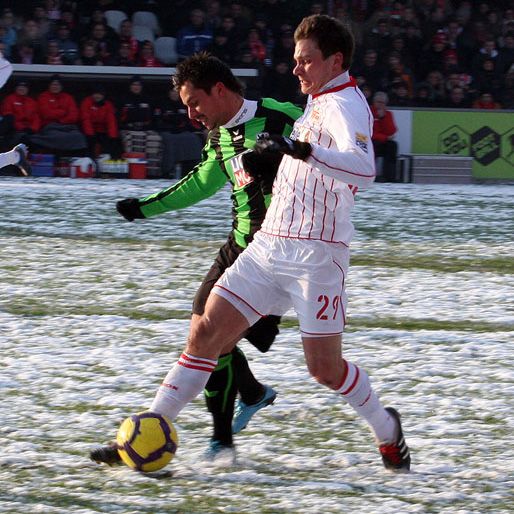 Zwei Fußballspieler im Wettkampf auf einem schneebedeckten Platz, einer mit grüner und der andere mit weißer Uniform.
