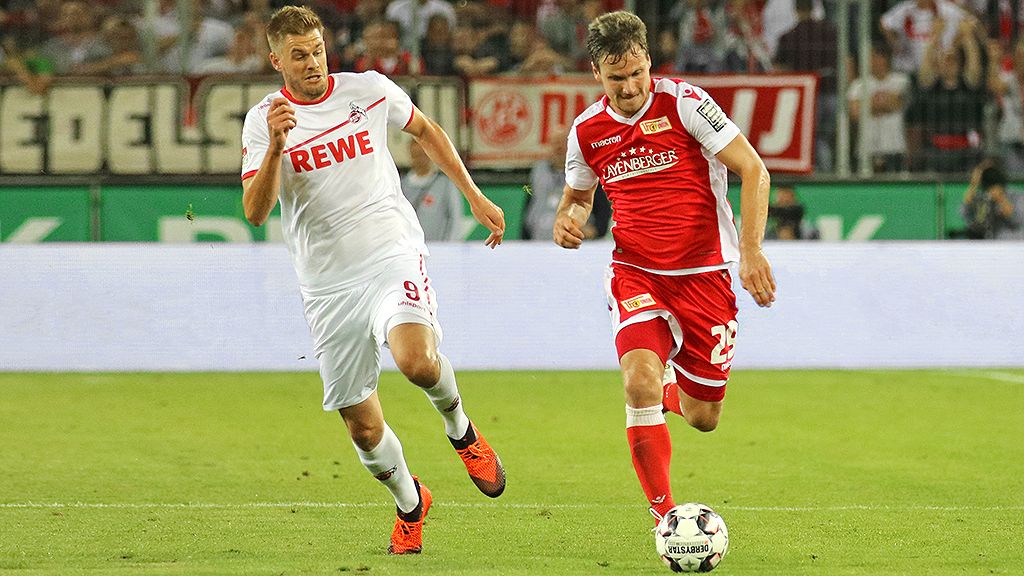 A player in a red jersey sprints with the ball while a player in a white jersey chases him, with a stadium atmosphere in the background.
