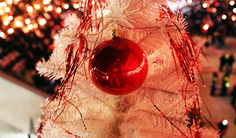 Weißer Weihnachtsbaum mit roten Ornamenten, im Hintergrund verschwommen Lichter und Menschenmengen.