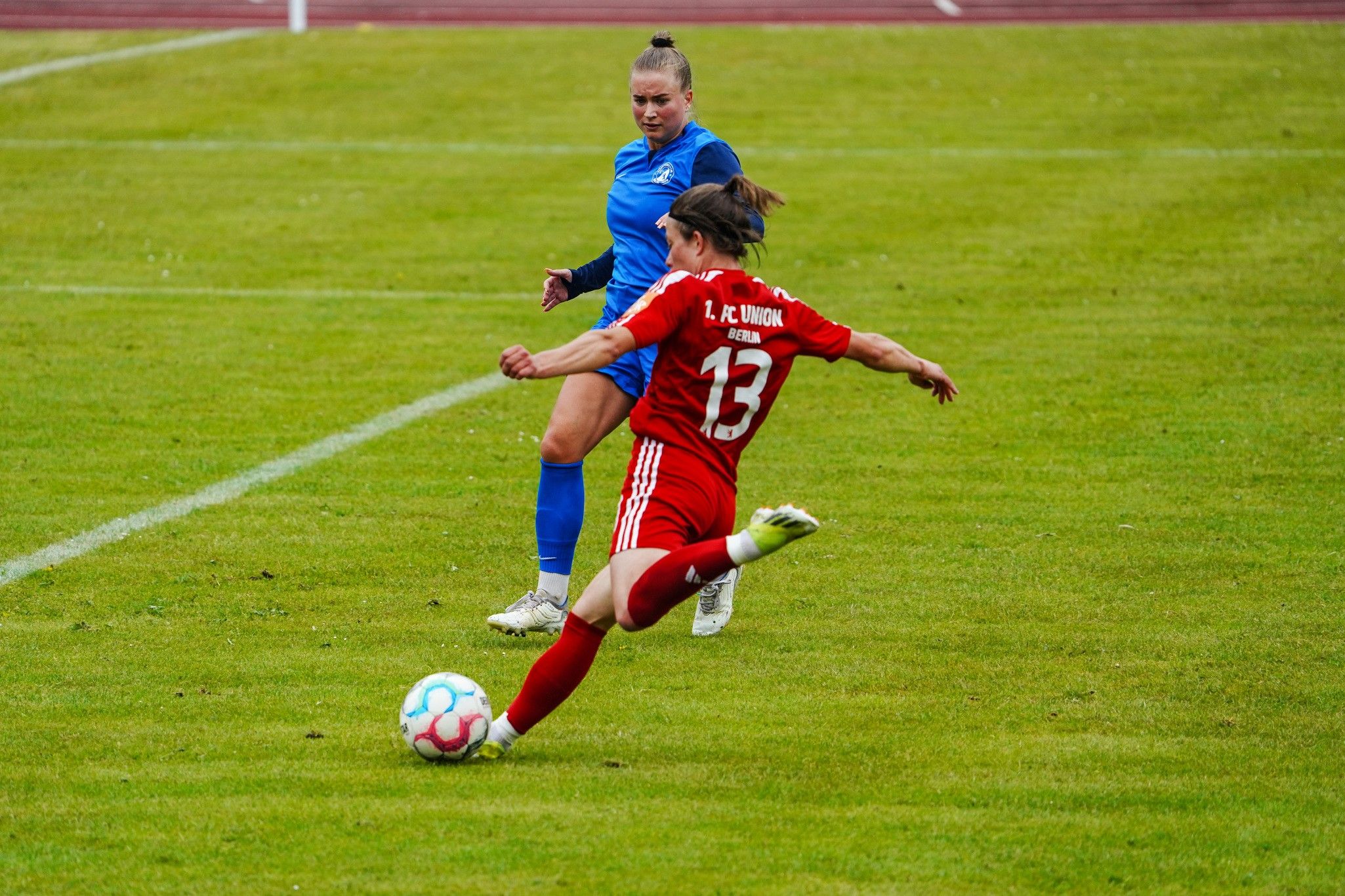 Eine Fußballspielerin in rotem Trikot schießt den Ball, während eine Torhüterin in Blau zuschaut.