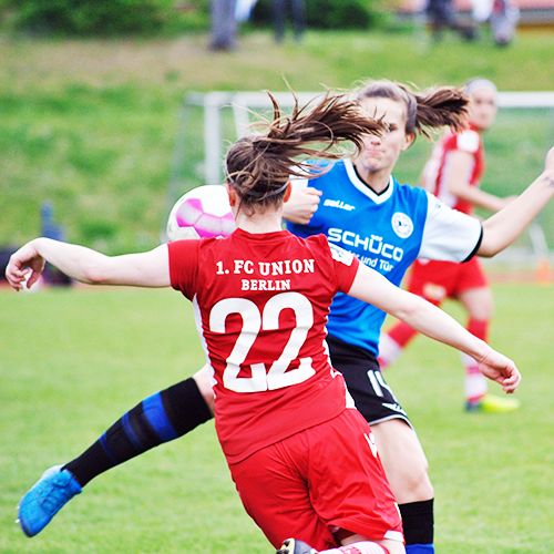Zwei Frauen spielen Fußball auf einem Rasenplatz, eine mit rotem Trikot und die andere mit blauem und schwarzem Trikot.