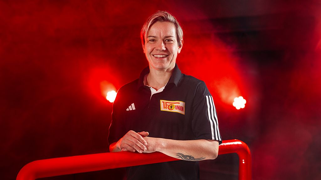 A smiling person in a black sports jersey with a logo stands against a red, smoky background.