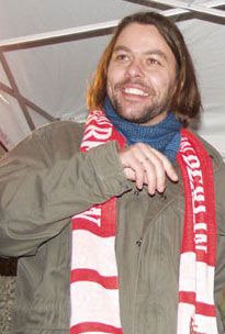 Mann mit langen Haaren trägt eine Jacke und einen roten Schal, lächelt und gestikuliert unter einem Zelt.