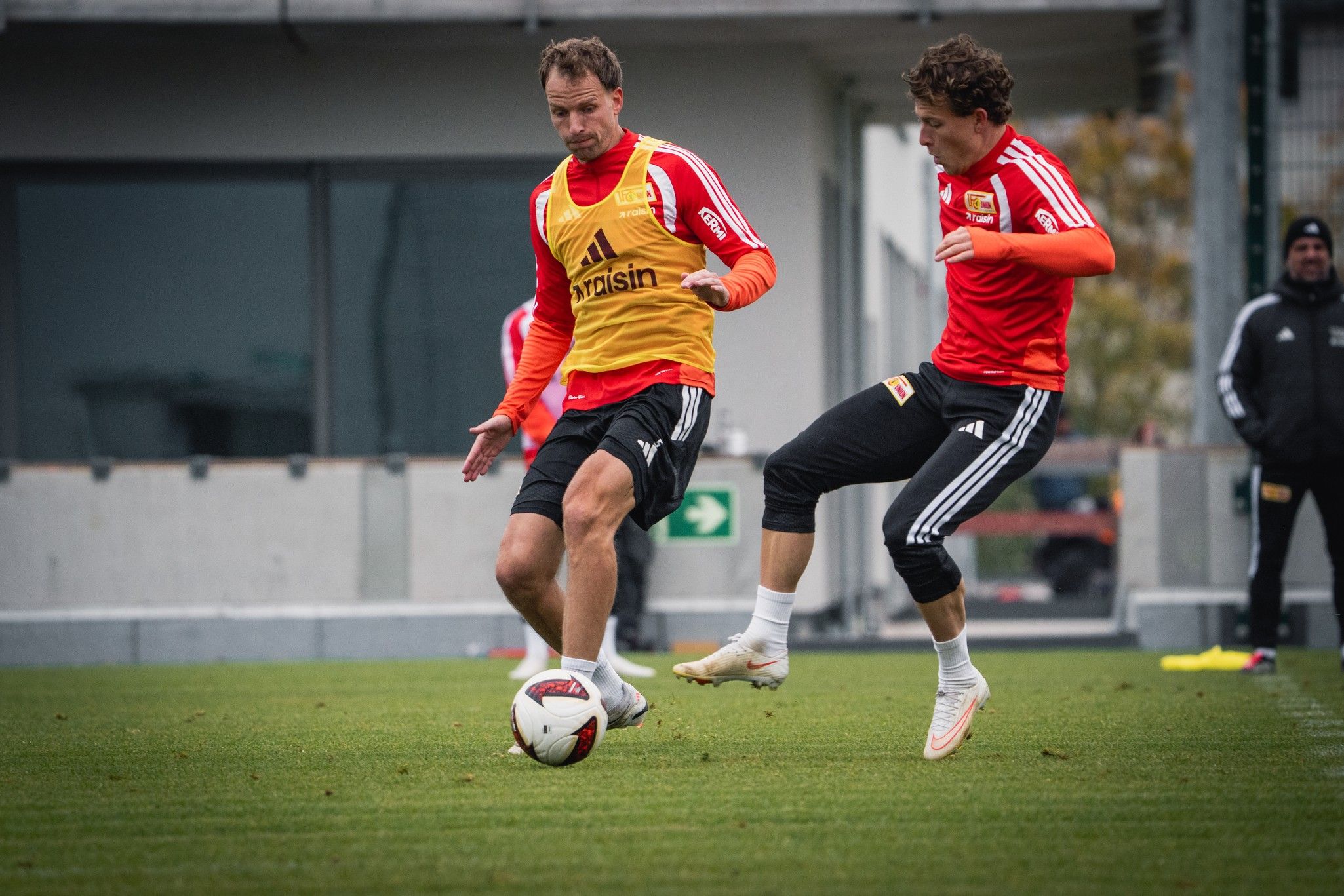 Zwei Fußballspieler in Trainingstrikots dribbeln und passen den Ball auf einem grünen Rasenfeld.